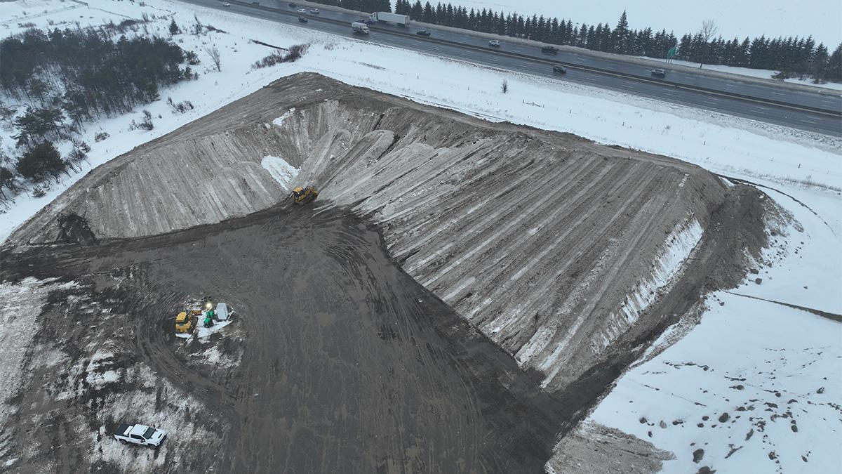 Construction vehicles near a large pile of snow at a storage site near a highway.