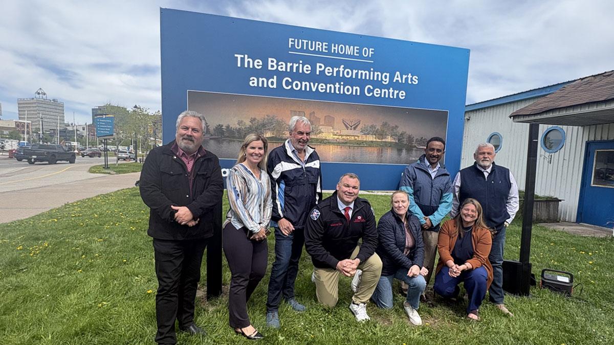 Group of people posing in front of a large sign with a rendering of the waterfront facility and text reading "Future home of the Barrie Performing Arts and Convention Centre".