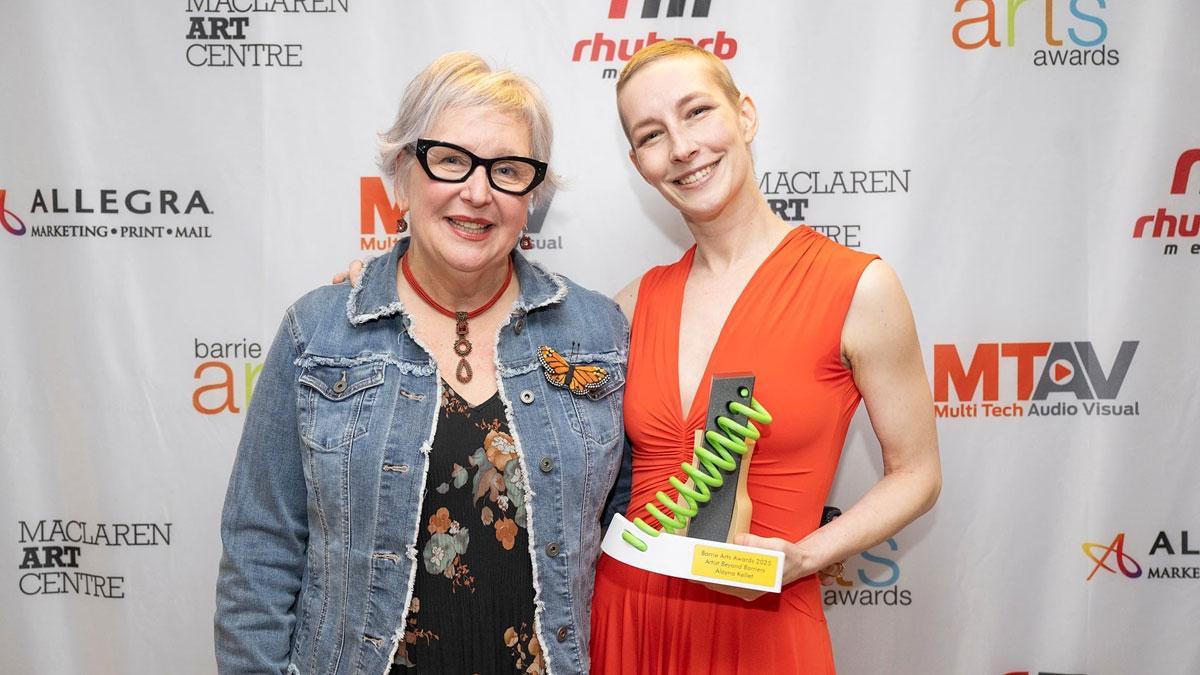 Two women pose together, one holding an award, against a decorated backdrop.