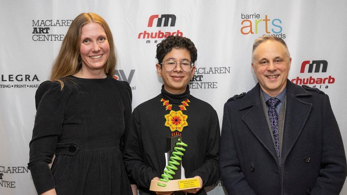 Three individuals pose together, smiling. The person in the middle holds an award.