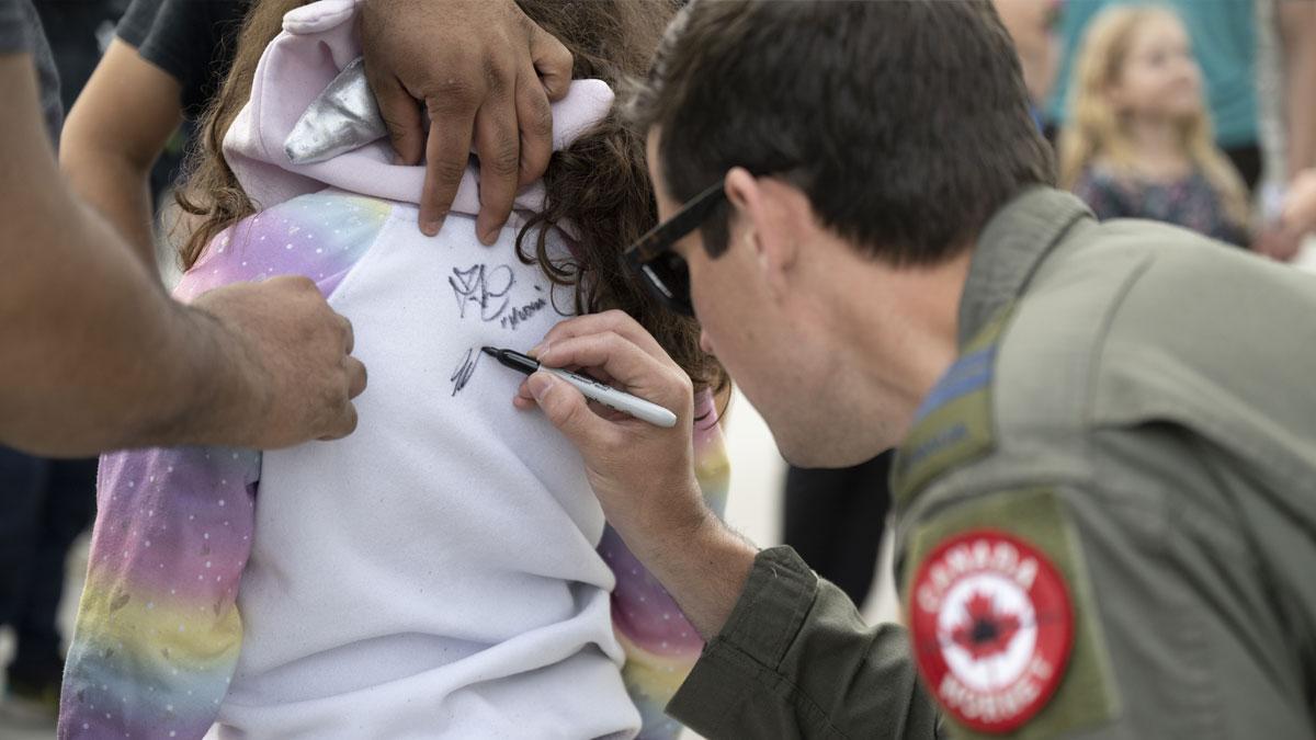 A person in a flight suit signs a child's hoodie while others watch in the background.