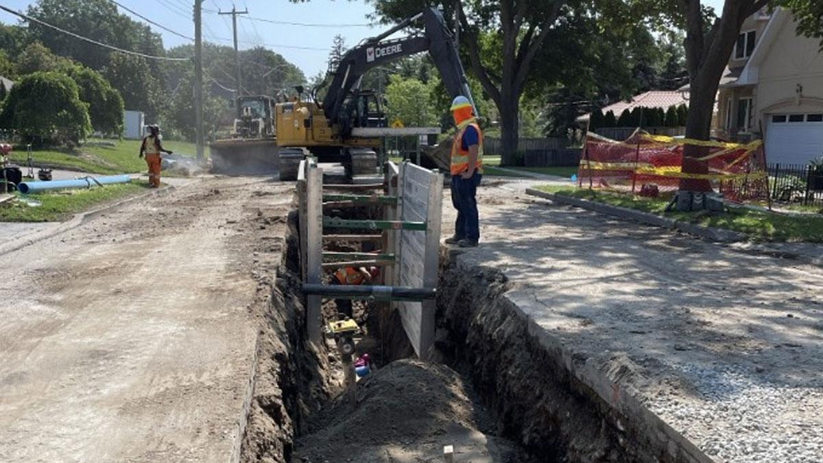 Construction workers operate machinery near a deep trench on a residential street.