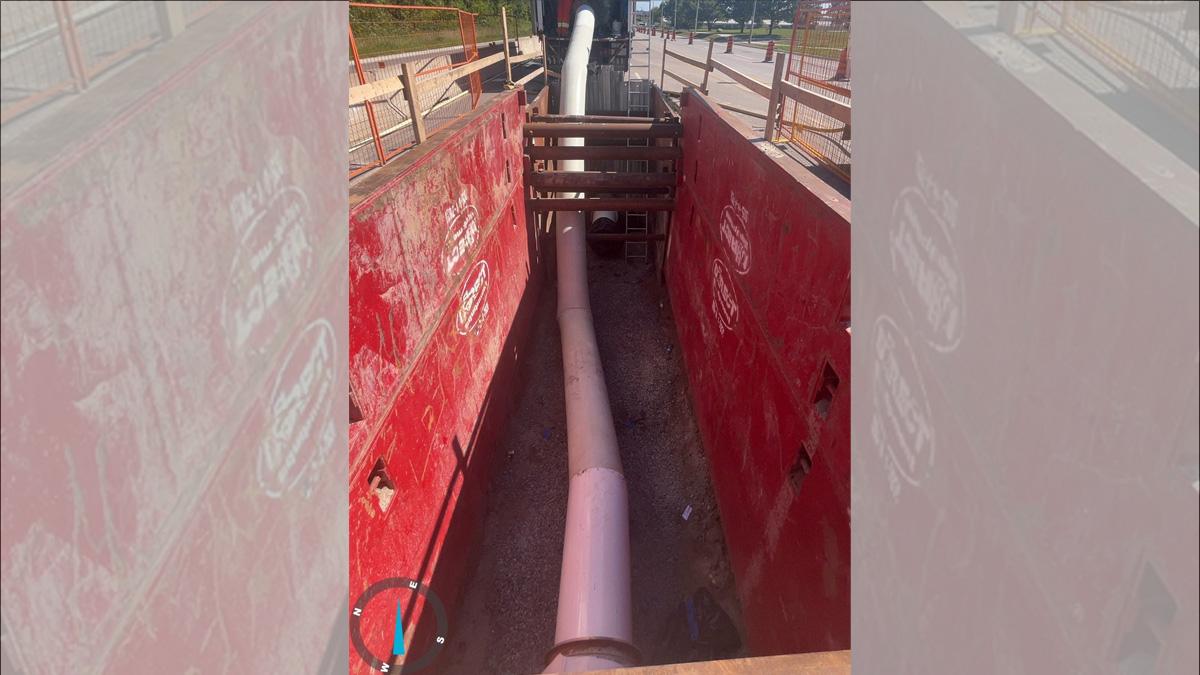 A construction site showing a dug out trench with a long white pipe reaching from the trench into the back of a truck.